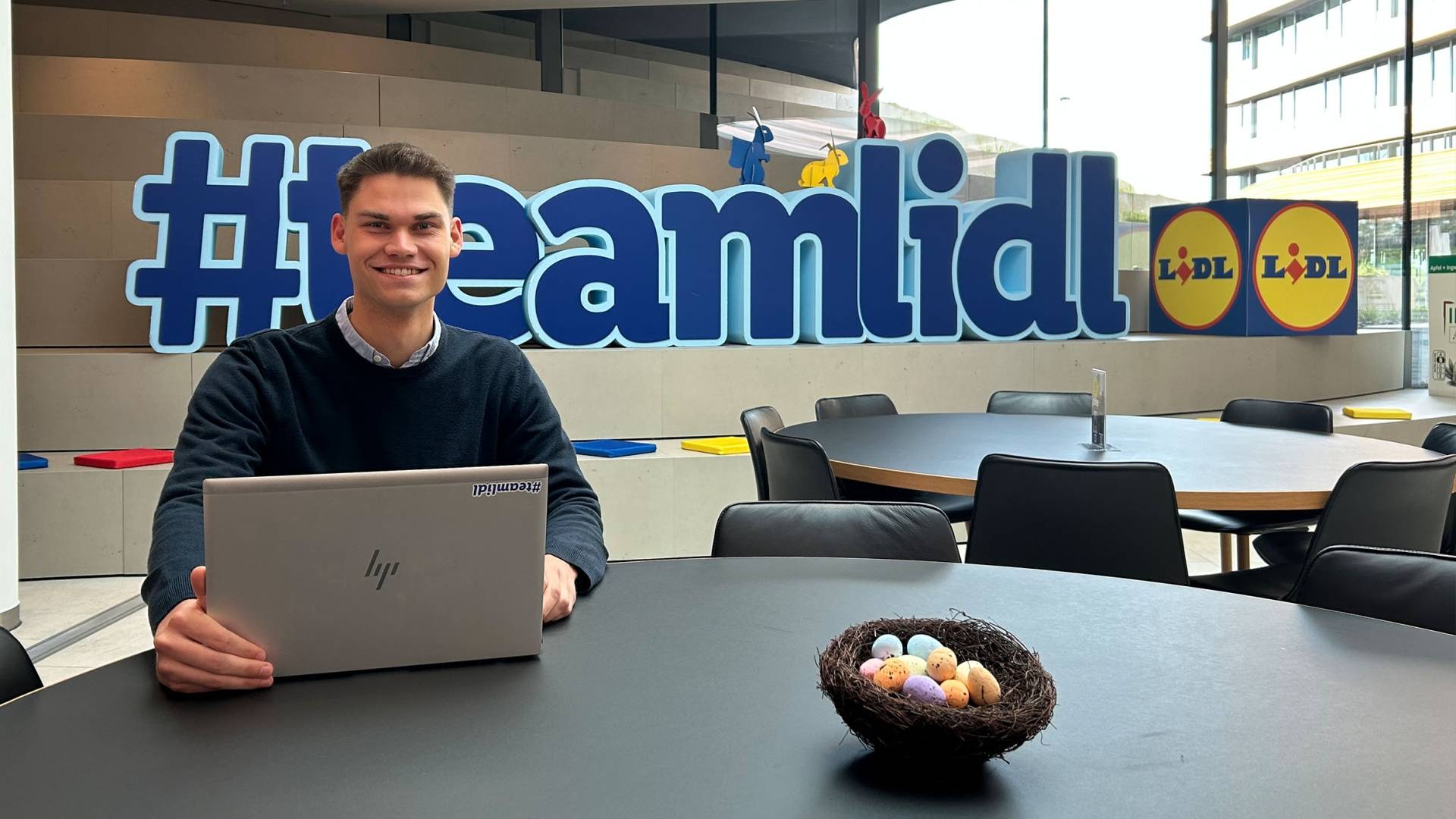 Nils sitzt vor dem #teamlidl Logo und arbeitet am Laptop.