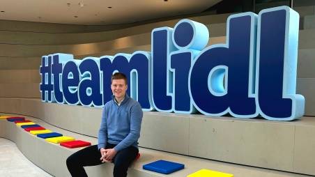 Maximilian sitzt vor dem #teamlidl Schild im Office in Bad Wimpfen.